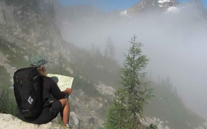 a person wearing a backpack sits on a rock and examines a map while fog obscures the mountains in the distance